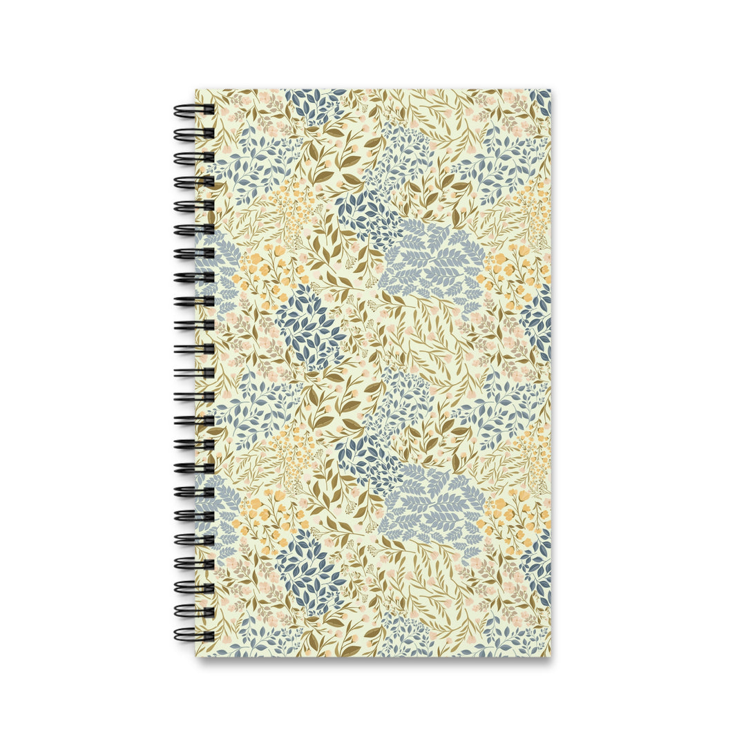 A spiral-bound notebook with a cream-colored cover decorated in a botanical pattern of blue, olive green, and mustard yellow leaves and flowers, shown on a plain white background.