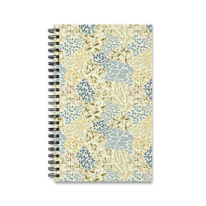 A spiral-bound notebook with a cream-colored cover decorated in a botanical pattern of blue, olive green, and mustard yellow leaves and flowers, shown on a plain white background.