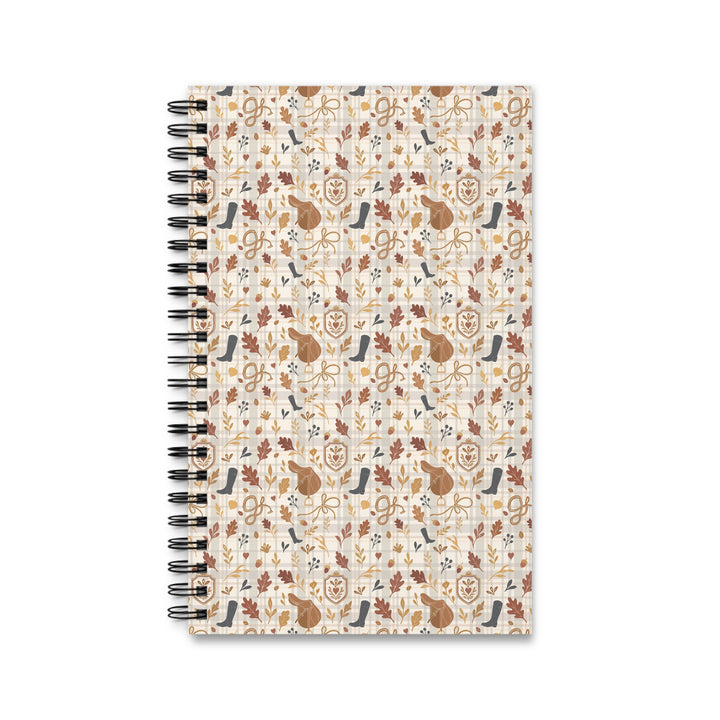 A spiral-bound notebook with a cream-colored cover featuring a repeating autumn-themed pattern of brown leaves, acorns, musical instruments, and birds, shown on a plain white background.