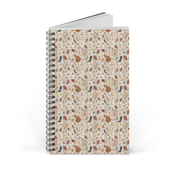 A spiral-bound notebook with a cream-colored cover decorated with a repeating pattern of autumn-themed illustrations, including leaves, birds, and animals in brown, orange, and blue tones. The notebook is shown standing upright on a clean white background.