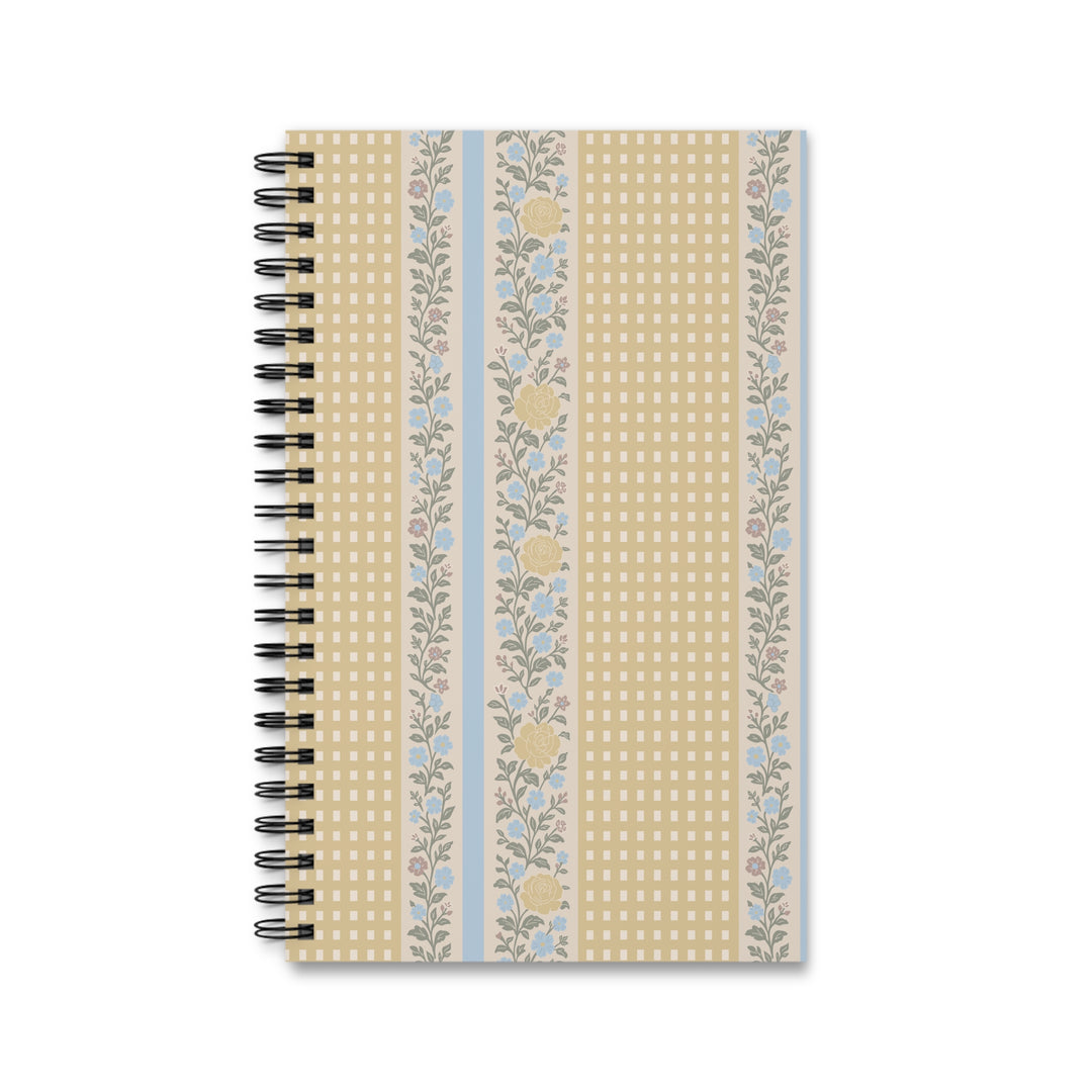 A spiral-bound notebook with a decorative cover featuring vertical stripes of beige gingham, floral patterns, and light blue accents, shown on a plain white background.