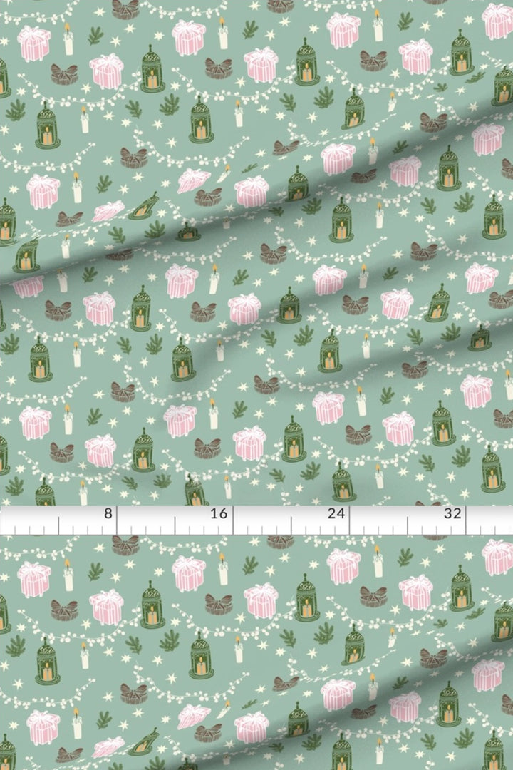 A close-up of patterned fabric with a repeating print of pink lanterns, green birdhouses, white candles, and small brown birds on a pale mint background, with a ruler across the middle showing scale.