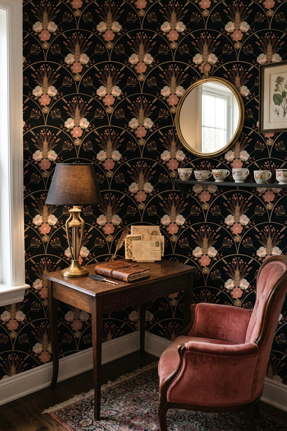 A styled interior corner featuring dark floral wallpaper with a repeating Art Deco fan pattern in black, blush pink, and beige. A dark wood writing desk with a brass lamp, books, and a mirror sits beside a dusty pink velvet armchair on a patterned rug.