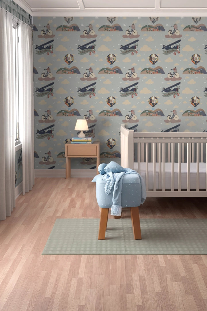 A styled nursery room featuring wallpaper with a vintage airplane and dog pattern on a light blue-gray background, a white wooden crib, a small wooden side table with a lamp, and a light blue stool with a matching throw blanket.