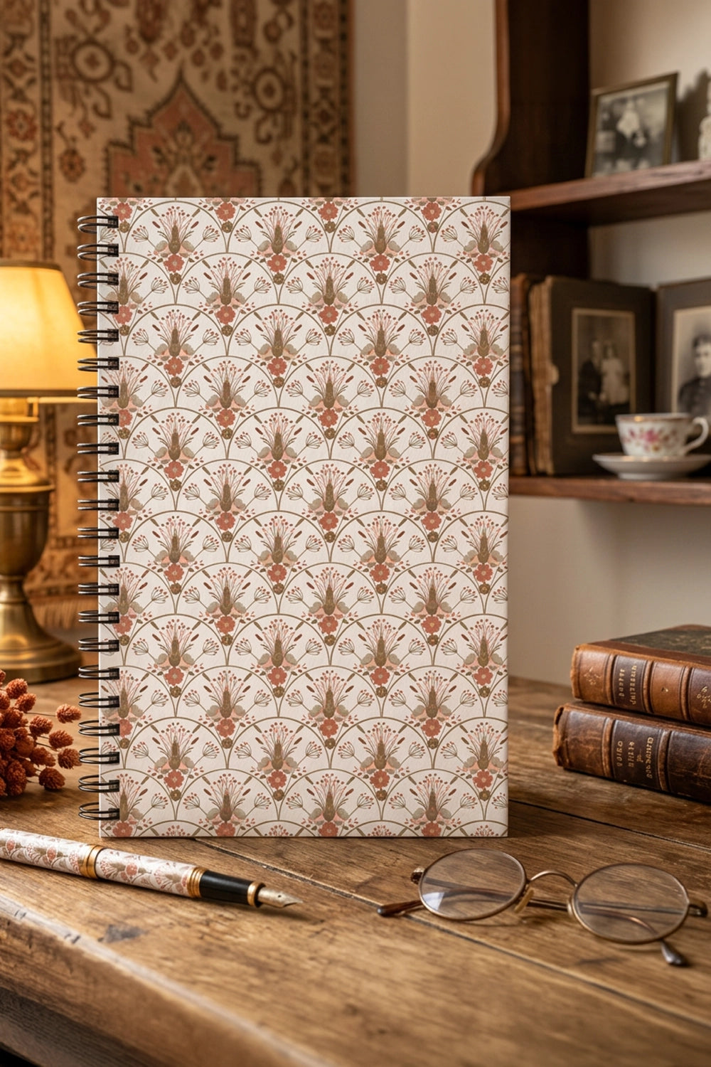 A spiral-bound notebook with a repeating ornate floral pattern in muted terracotta and brown on an off-white background, standing on a rustic wooden table with a pen, eyeglasses, and books nearby.