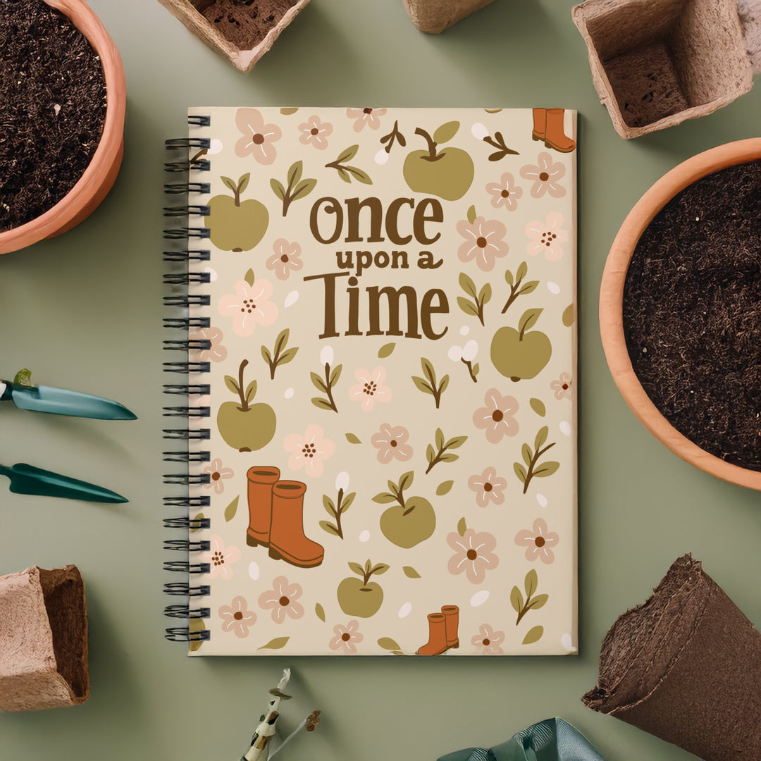 A spiral-bound notebook with an autumn-themed cover design reading 'once upon a time,' decorated with illustrated apples, flowers, leaves, boots, and a small tree on a beige background. The notebook is photographed from above on a muted sage green surface, surrounded by gardening props including pots of soil, seed-starting bags, and small tools.