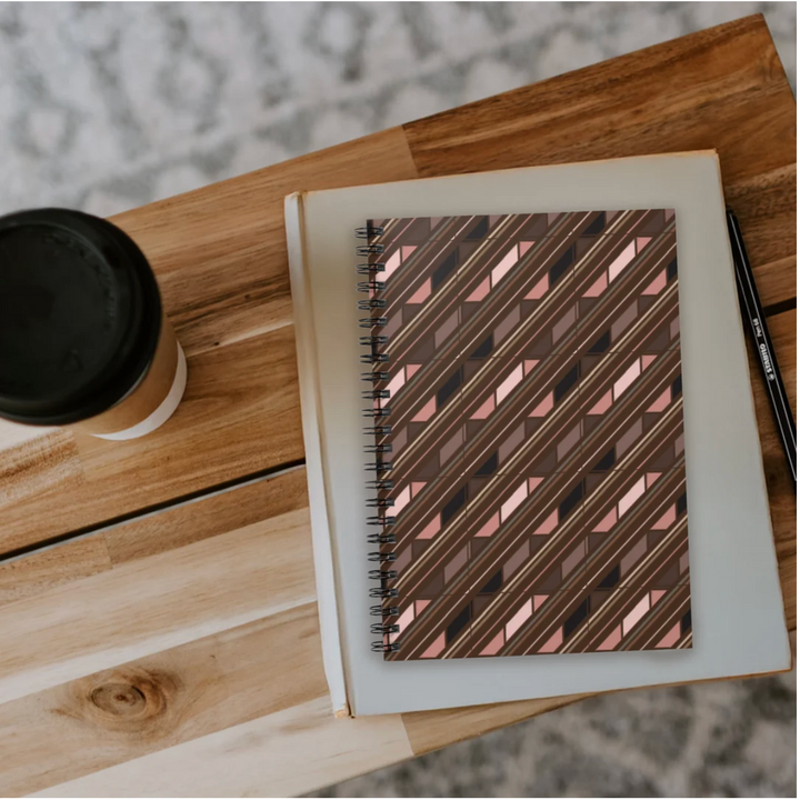 A spiral-bound notebook with a metallic rose gold and brown geometric patterned cover, resting on a light wood table next to a black coffee cup and a black pen.