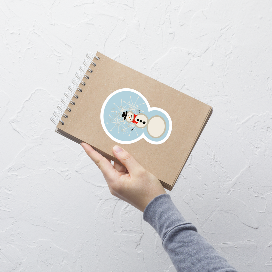 display of a snowman sticker that shows him in a red scarf and button down coal on his chest.  Snowflakes are also falling behind him. It's shown displayed on a notebook front
