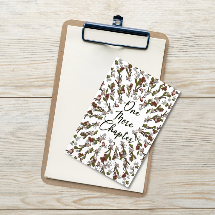 A bookmark with the words "one more chapter" written on it with flowers surrounding it 