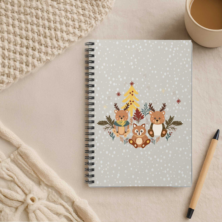 A spiral-bound notebook with a winter forest cover design featuring three bears, a Christmas tree, and snowflakes, styled on a beige surface with a knit blanket, a cup of coffee, and a pen.