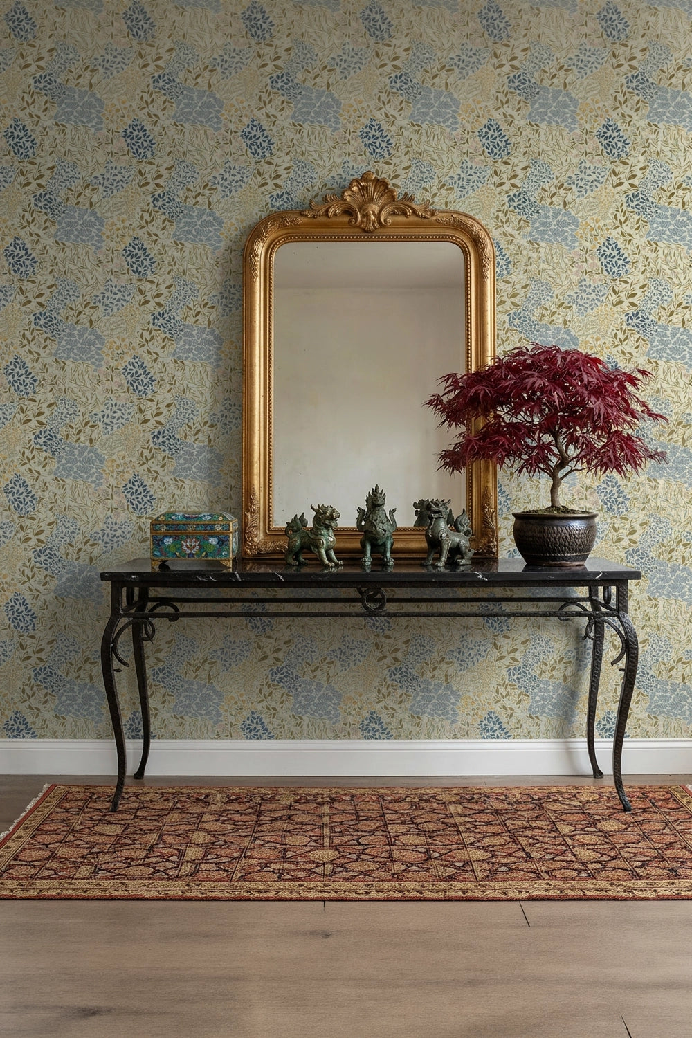 An ornate gold-framed mirror hangs above a black console table against a blue and beige floral wallpapered wall. A potted red-leafed tree and small decorative figurines sit on the table, with a patterned rug on the floor.