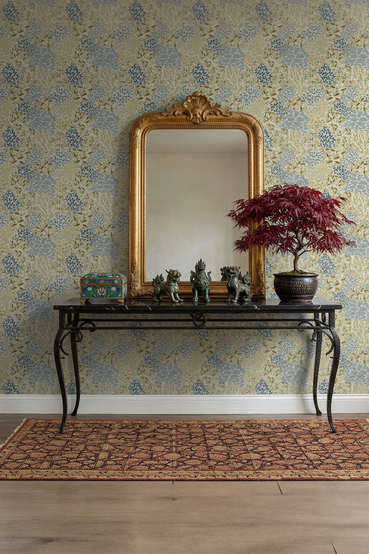 An ornate gold-framed mirror hangs above a black console table against a blue and beige floral wallpapered wall. A potted red-leafed tree and small decorative figurines sit on the table, with a patterned rug on the floor.