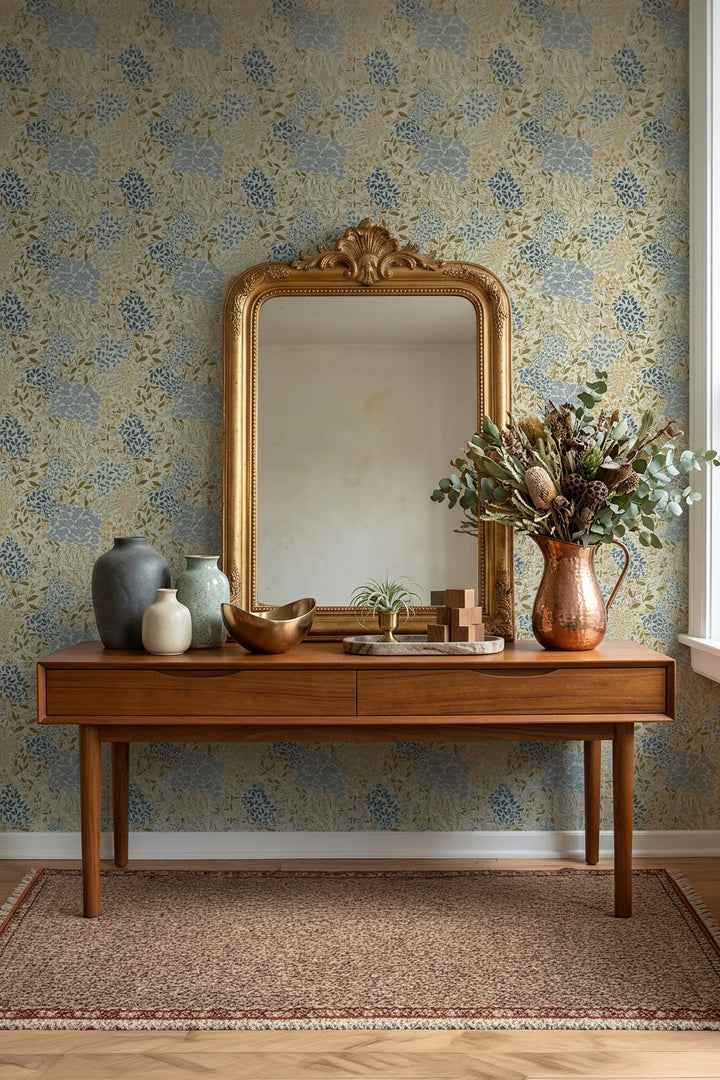 A styled interior scene featuring a large ornate gold-framed mirror leaning against a blue and beige floral patterned wallpaper wall above a mid-century modern wooden console table. The table is decorated with ceramic vases, a copper-toned vase of dried flowers, and a small tray, set on a textured area rug.