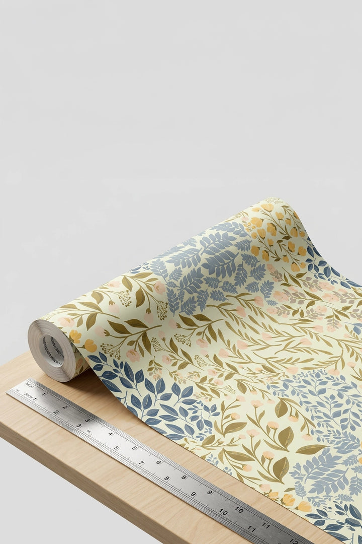 A roll of wallpaper with a botanical leaf pattern in muted blue, olive green, and soft peach on a cream background, shown partially unrolled on a light wood surface with a metal ruler for scale.